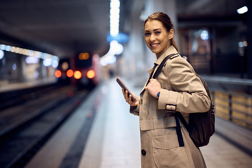 Young happy woman using smart phone while for train at subway and looking at camera.