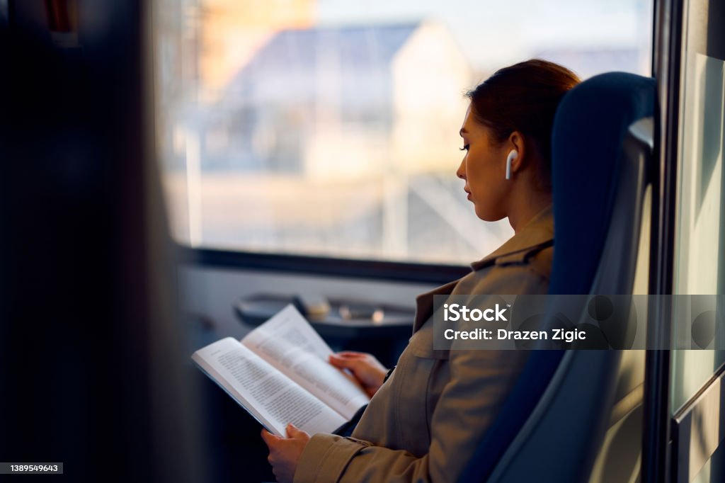 Young female passenger reading book while traveling by train. Young woman reading a novel while listening music over earbuds and commuting by train. Train - Vehicle Stock Photo