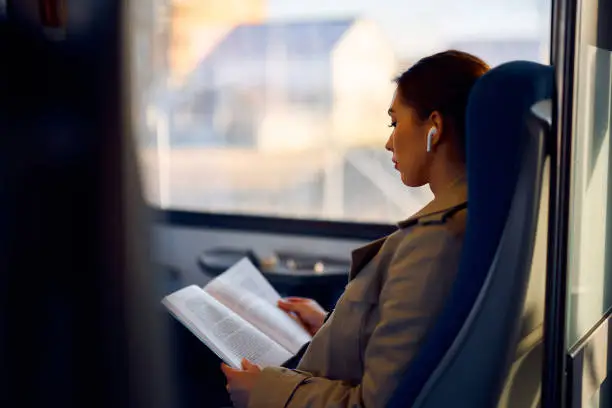 Young woman reading a novel while listening music over earbuds and commuting by train.