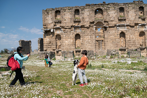 Photo of 8,5 years old boy playing with pet dog while mother is photographing them via smartphone. Grandmother and cousin are seen on the background in old ruins of Aspendos. Shot under day light.