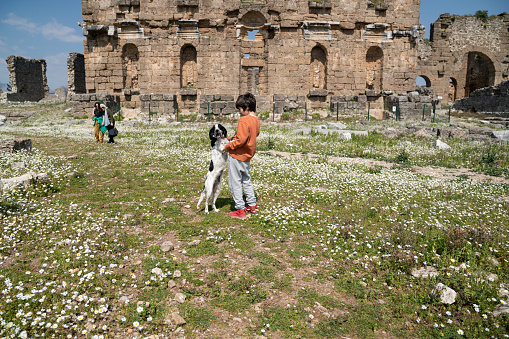 Photo of 8,5  years old boy playing with pet dog in old ruins of Aspendos during springtime. Old ruins of basilica, grandmother and cousin are seen on the background. Shot under daylight.