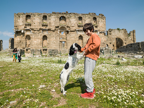 Photo of 8,5  years old boy playing with pet dog in old ruins of Aspendos during springtime. Old ruins of basilica, grandmother and cousin are seen on the background. Shot under daylight.