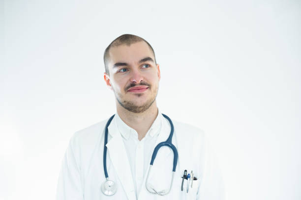 Portrait of a male doctor with stethoscope looking away and thinking. Medical professional standing against white wall looking at copy space.