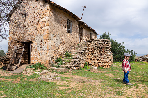 Photo of 3 years old boy in old countryside stone village house. He is wearing a hat and a plaid shirt. Shot under daylight. The house is stone made traditional Turkish style village house and is abandoned.