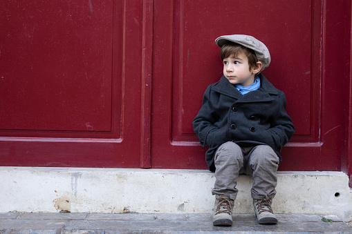 Portrait of preschooler boy wearing a fashionable coat, hat and scarf in outdoor. He is sitting in front of an old antique wooden door.