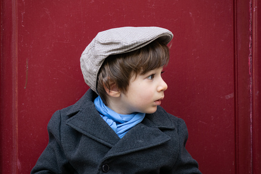 Portrait of preschooler boy wearing a fashionable coat, hat and scarf in outdoor. He is standing in front of an old antique wooden door.