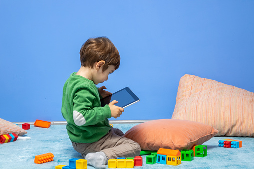 Photo of 3,5 years old preschooler boy using digital tablet while multi colored toy blocks are seen around. He is wearing a green sweater and brown pants and sitting on carpet in living room. Shot indoor.