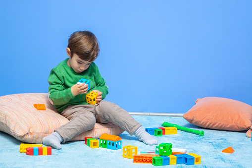 Photo of 3,5 years old preschooler boy playing with multi colored toy blocks. He is wearing a green sweater and brown pants and sitting on carpet in living room. Shot indoor.