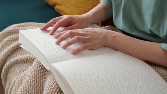 Blind woman reading braille book sitting on sofa, touching letters on sheet of paper using his fingers, poorly seeing person learning to read, concept of home education for people with disabilities