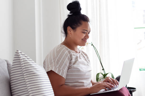 Happy young woman working from home Cheerful millennial age woman of Pacific Islander descent sits on the sofa at home and smiles while working on a laptop computer. pacific islander ethnicity stock pictures, royalty-free photos & images