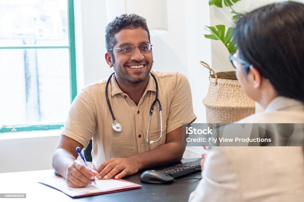 Cheerful male doctor meeting with a patient A cheerful male doctor of Indian descent sits at a desk in his office and takes notes while meeting with a female patient. Doctor Stock Photo