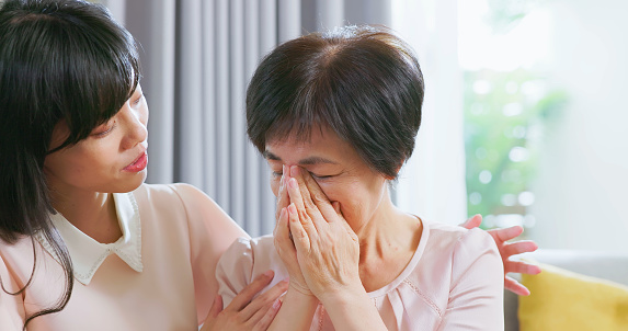 asian worried young daughter is comforting her sad senior mother and apologizing to her with hug in living room at home