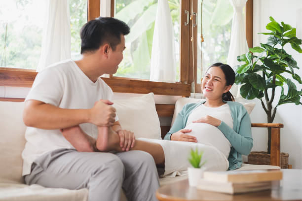 Asian young husband massaging pregnant wife feet at home, happy family stock photo