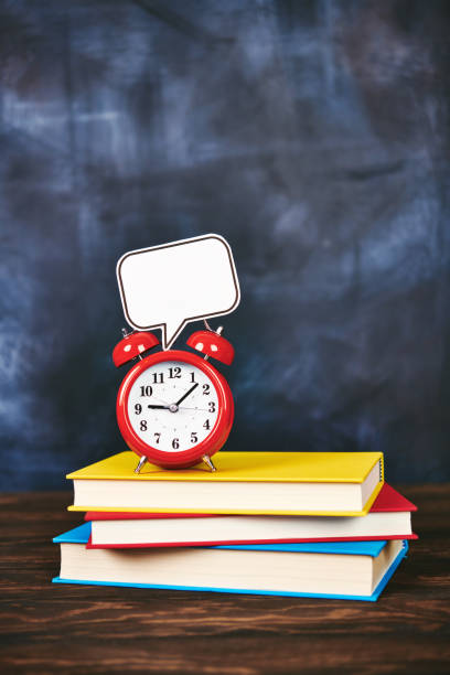 Alarm clock with empty speech bubble for message on top of a stack of school books in front of a chalkboard