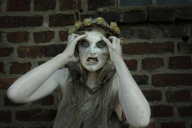 A teenage girl dressed as a Halloween horror figure. She clutches her head and her face which are filled with horror and pain. She wears a thorny floral crown on her head filthyrags as clothes.