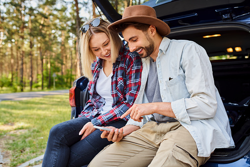 Young couple sitting in car trunk looking at smartphone before going hiking