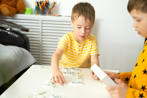 Boys play dominoes at home on the table. Older brother teaches younger brother to play dominoes. Concept of inclusive education and early development