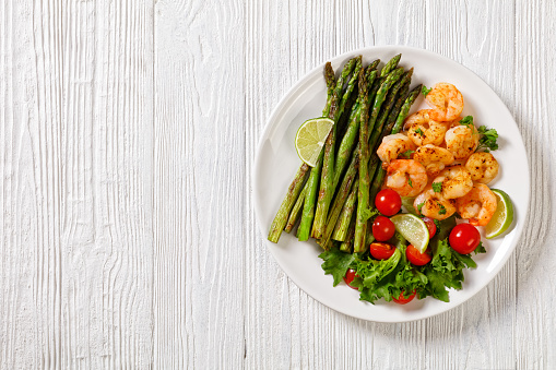 toasty asparagus with fried prawns, lime, lettuce and cherry tomatoes on a white plate on wooden table,  flat lay, close-up, free space
