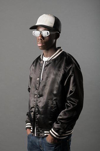 Confident African American male rapper in cap and sunglasses standing with hands on pockets on gray background in light studio