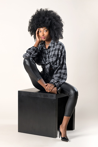 Beautiful slender Black woman in stylish leather pants and high heels sitting with hand to chin in a pensive mood on a black wooden stool in a studio portrait