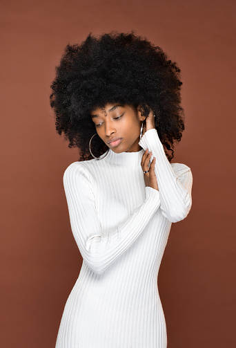 Slender calm Dominican lady with Afro hairstyle in white dress touching hair and looking down against brown background