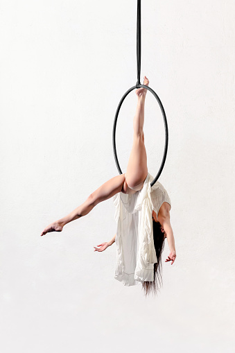 Full body side view of flexible slim barefoot acrobatic female exercising on aerial hoop against white background in light studio