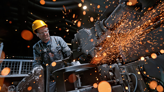 Male worker producing stream of hot steel sparks while grinding product.