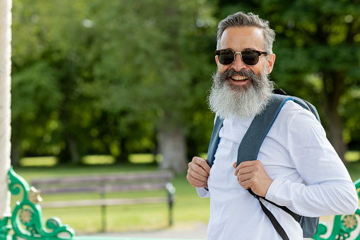 A portrait of a mature man wearing a backpack and sunglasses, standing outdoors in Newcastle upon Tyne, England. He is looking at the camera and smiling.