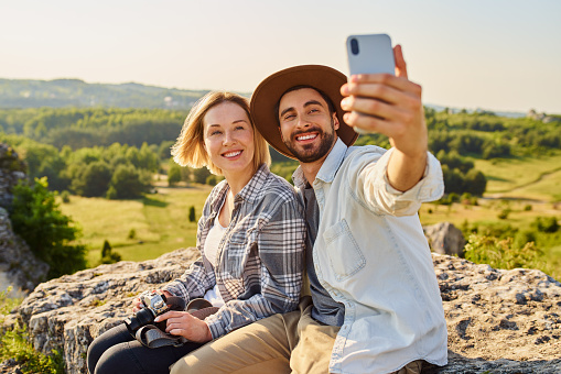 Smiling young couple taking selfie while sitting on rock in mountains during summer hike
