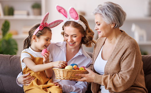 Happy multi generational family grandmother mother and daughter in bunny ears laugh and play with eggs at home in a cozy living room during preparation for   Easter holiday