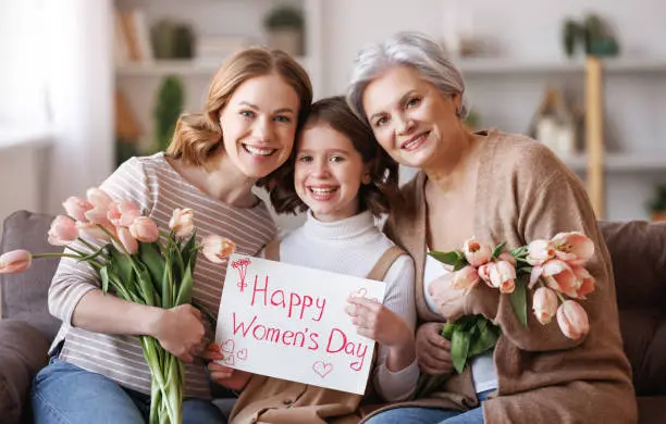 Happy International Women's Day! Smiling multi-generational family grandmother, mother and daughter with flowers and  poster cheerfully celebrate the spring holiday Mother's Day at home