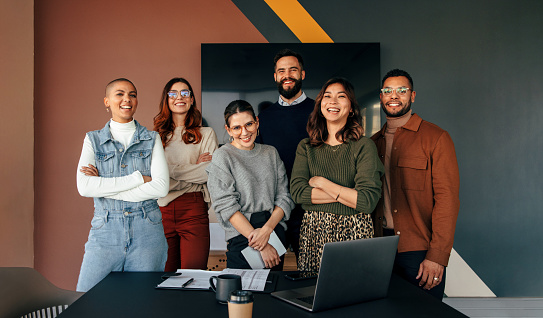 Multicultural business team smiling at the camera while standing behind a table in a boardroom. Group of successful young businesspeople working together in a modern workplace.