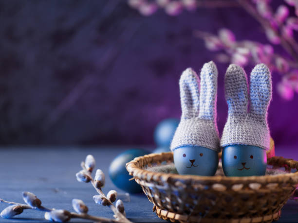 istock blue eggs in crocheted hats with bunny ears and painted muzzles in a wicker basket, willow branches. neon background with copy space 1389653566