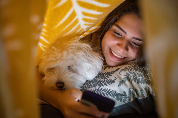 istock Joyful teenage girl, using her mobile phone, while lying under the bed sheet with her dog 1389656034