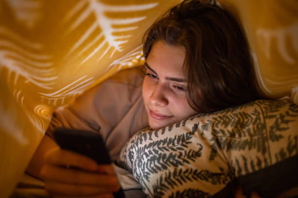 adolescente caucasiana, usando seu celular sob o lençol - one teenage girl only on the phone bed lying on front - fotografias e filmes do acervo