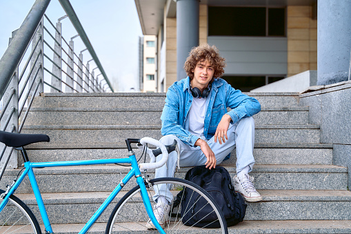 Full body of positive curly haired male in casual clothes with headphones on neck sitting on staircase near bike for riding on city streets