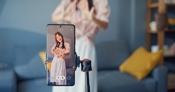 Happy young Asian girl blogger front of phone camera record video enjoy with dance content in living room at home. Social distance coronavirus pandemic concept. Freedom and active lifestyle.