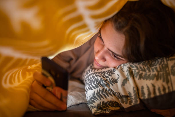 adolescente, usando seu celular sob o lençol - one teenage girl only on the phone bed lying on front - fotografias e filmes do acervo