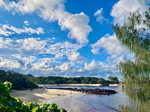 Horizontal seascape of calm beach waters with promenade rocks and tropical trees u der a blue cloudscape sky at Torakina Beach Brunswick Heads near Byron Bay NSW Australia