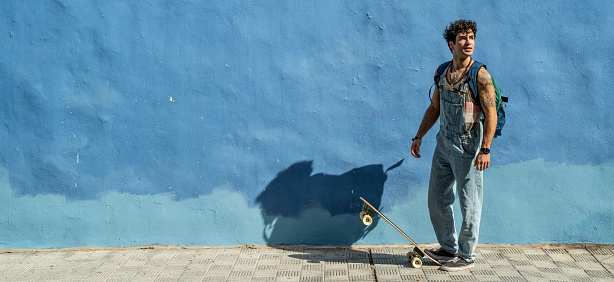 Young handsome skater man with backpack and waveboard. Sunny summer day in the city. Real people lifestyle. Hobby concept.