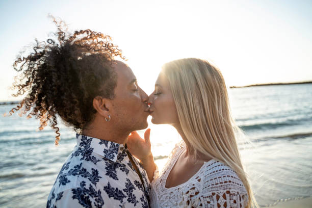 pareja multiétnica besándose, divirtiéndose juntos, saliendo en la playa durante la puesta de sol. gente enamorada. luna de miel. emociones reales - honeymoon beach couple heterosexual couple fotografías e imágenes de stock