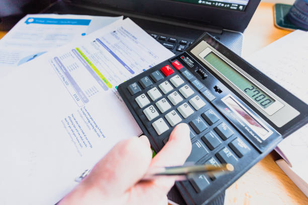 Portrait of a senior caucasian man in his 60s checking his energy bills at home. He has a worried expression and touches his face with his hand while looking at the bills. Focus on the man while the interior architecture of the house is defocused.