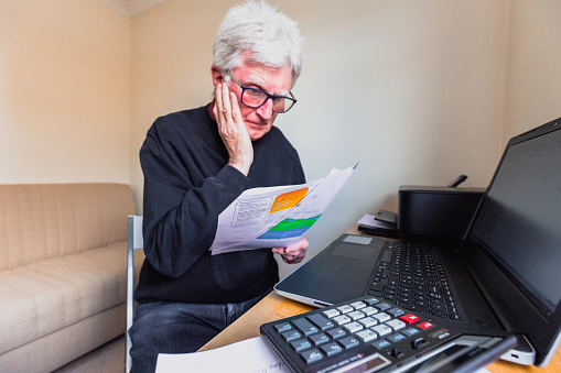 Portrait of a senior caucasian man in his 60s checking his energy bills at home. He has a worried expression and touches his face with his hand while looking at the bills. Focus on the man while the interior architecture of the house is defocused.