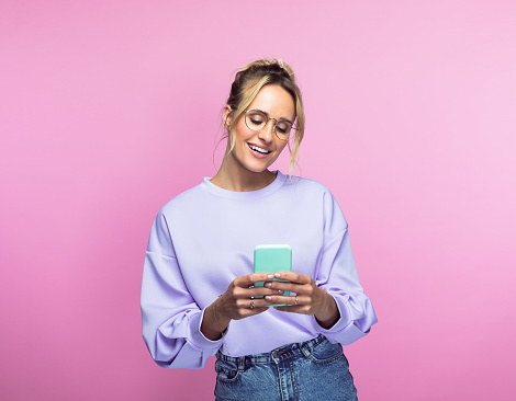 Happy beautiful woman using smart phone while standing against pink background.