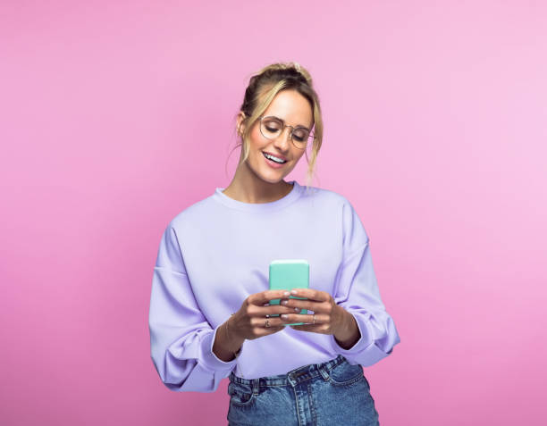 Happy woman using smart phone Happy beautiful woman using smart phone while standing against pink background. mobile phone text messaging telephone women stock pictures, royalty-free photos & images