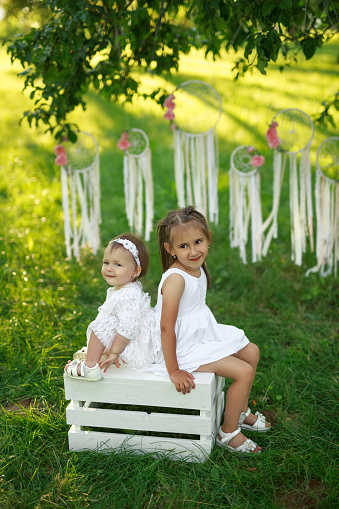 Two little sisters in white dresses are sitting on a box in the garden. Dream catchers with flowers hang on the trees.
