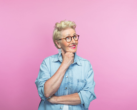 Thoughtful senior woman with hand on chin smiling against pink background