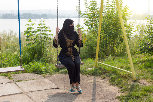 A Muslim woman in national clothes rides on a swing on the riverbank enjoying the fresh air.