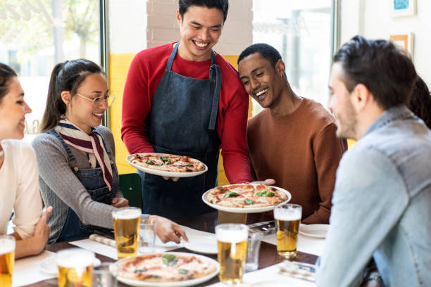 serveur confiant servant de délicieuses pizzas margherita à des amis multiculturels dans un restaurant pizzeria confortable - amis multiethniques s’amusant ensemble à la pizzeria en mangeant de la pizza et en buvant de la bière blonde - pizza pizzeria restaurant waiter photos et images de collection