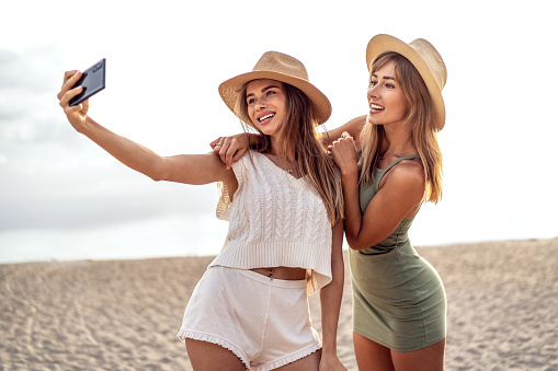 Two happy blonde caucasian sisters having fun on the beach during summer, taking selfie with mobile phone, smiling and looking to the smartphone camera. Beautiful girls wearing hats. Friendship. Real people emotions.